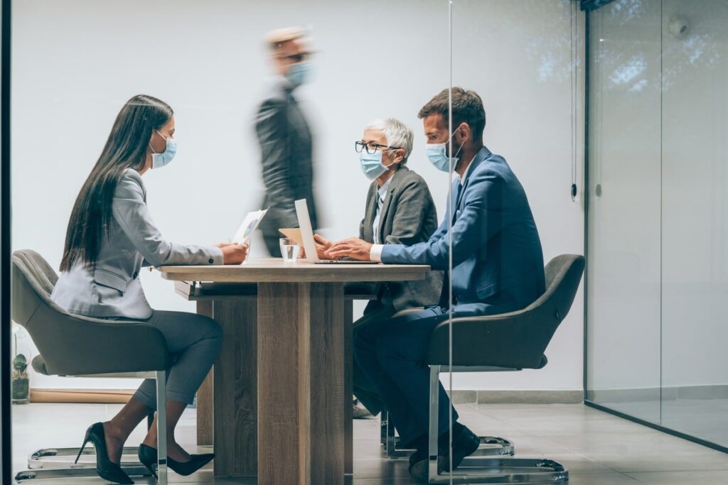 Shot of a group of business people with protective face masks having a meeting in board room. Businessman walking in blurred motion around his colleagues. Group of entrepreneurs wearing masks and sitting at a distance in the office. Business people wearing masks in the office for safety during pandemic situation. Corporate business team on meeting in the office during Coronavirus/COVID-19 pandemic. The view is through glass.