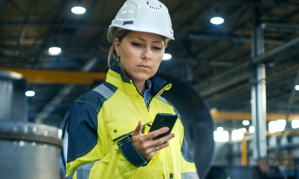 Female Industrial Worker In The Hard Hat Uses Mobile Phone While Walking Through Heavy Industry Manufacturing Factory. In The Background Various Metalwork Project Parts Lying