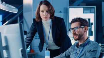 Futuristic Machine Engine Development Engineer Working On Computer At His Desk, Talks With Female Project Manager. Team Of Professionals Working In The Modern Industrial Design Institution